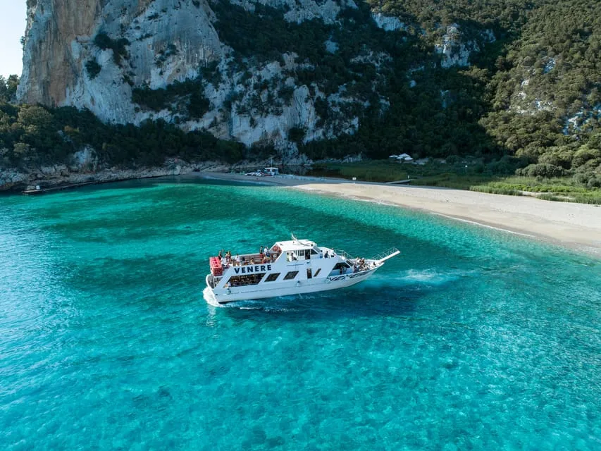 A white tour boat in turquoise water off Cala Luna beach, with the cliff and its caves behind